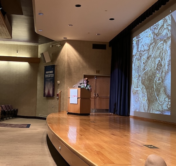 Nat Clayton presents her keynote talk on stage. Behind her is a slide showing a videogame map. Justin Bortnick is slightly visible in the foreground.