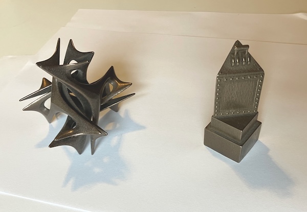 Two bronze-colored metal objects. On the left is a curved geometric abstraction with tetrahedral symmetry. On the right is a Myst marker switch. Each is about three inches high.