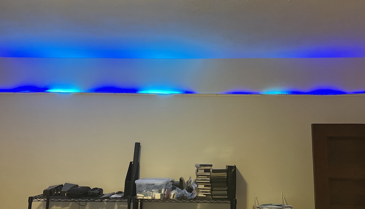 An LED strip runs along the top of a wall, above some wire-rack shelves of CD-ROMs and assorted junk. The strip glows in irregular segments of deep blue and bright cyan. The colors are bright enough to blow out the image somewhat; they shine up the wall and reflect off the ceiling.