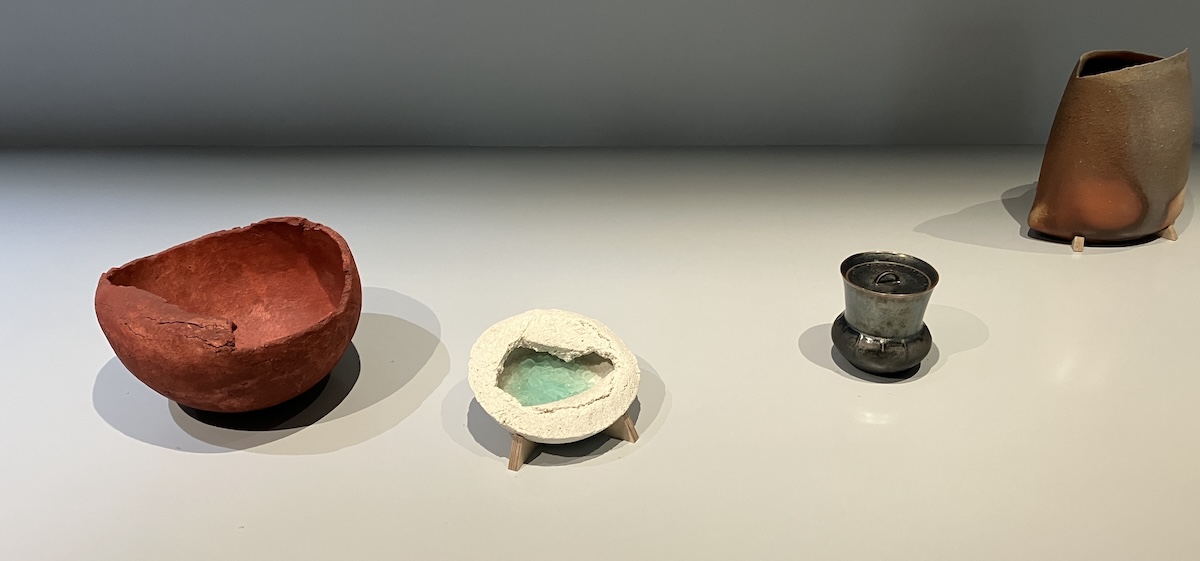 Four diverse clay vessels. The first is brick-red, bowl-shaped, rough in outline. The second is smaller, off-white, with an almost bread-like texture and a pool of glassy, cracked glaze inside. The third is a lidded urn with a dark and very metallic glaze. The fourth is tall and irregular with a pattern scorched in red and black into the surface.