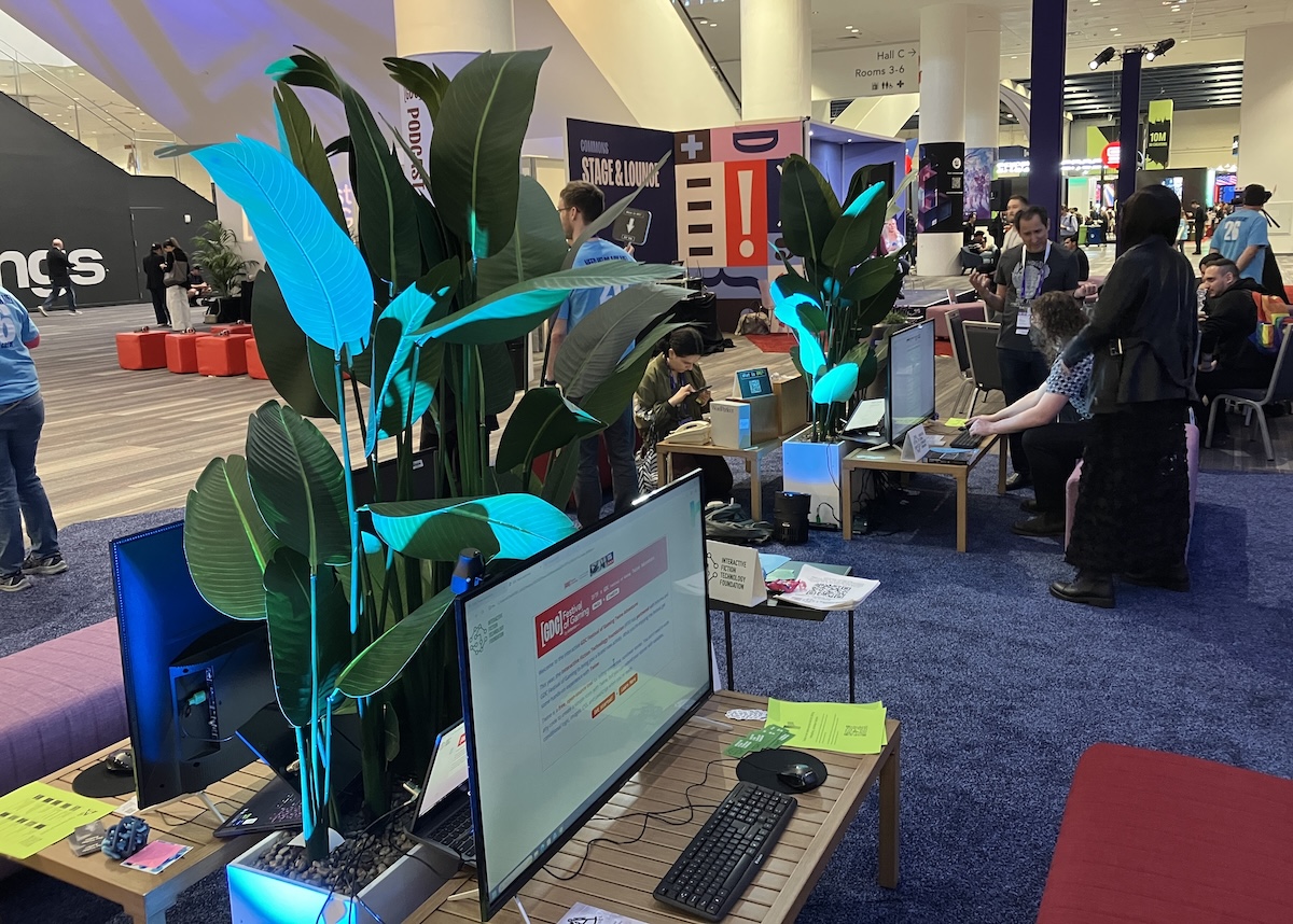 The expo hall at the Moscone Center. Large monitors are set up on low tables with benches. The nearest shows the opening screen of a Twine game. Farther away, someone is filling out a survey form; two other people look on.