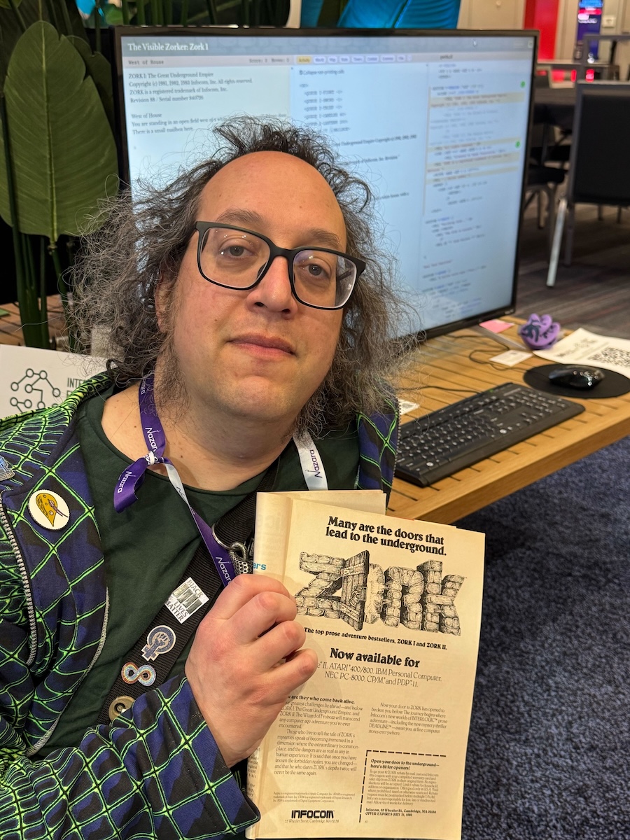 Me holding a magazine ad for _Zork 1_ and _Zork 2_. The headline "Many are the doors that lead to the underground" stands above the familiar Zork opening-door logo. "Now available for Apple II, ATARI 400/800, IBM Personal Computer, NEC PC-8000, CP/M, and PDP-11." In the background is a screen displaying Visible Zork 1.
