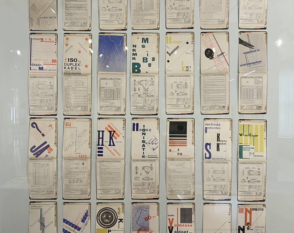 A wall display of pages from technical manuals. The bottom of each page is a technical blueprint, but the top is a startling assortment of Dutch words and letters laid out in varying fonts, angles, and colors.